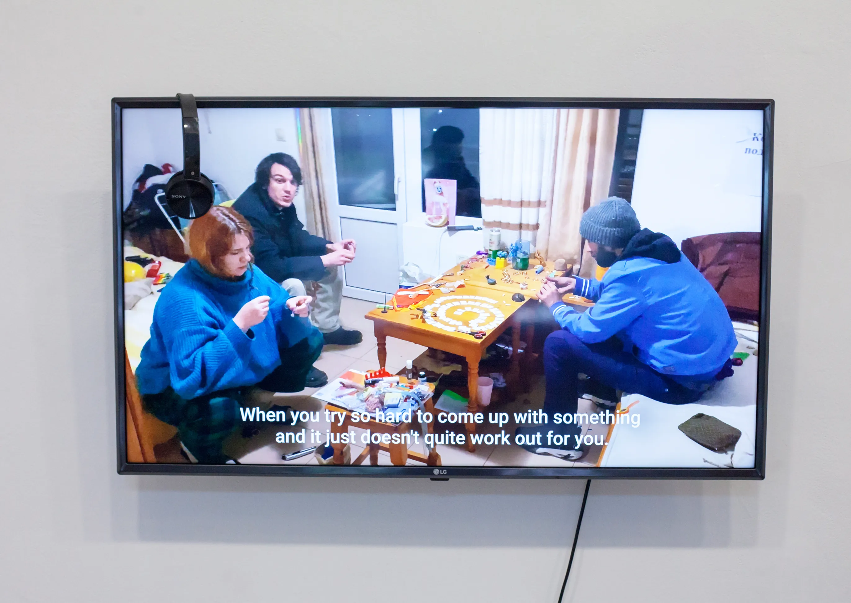 Video still on a wall-mounted screen showing three people around a table, subtitled 'When you try so hard to come up with something and it just doesn't quite work out for you.'