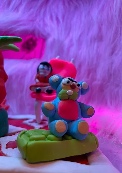 Detail of a blue clay bear figure seated on a green cushion, with a small figure in a red chair behind it