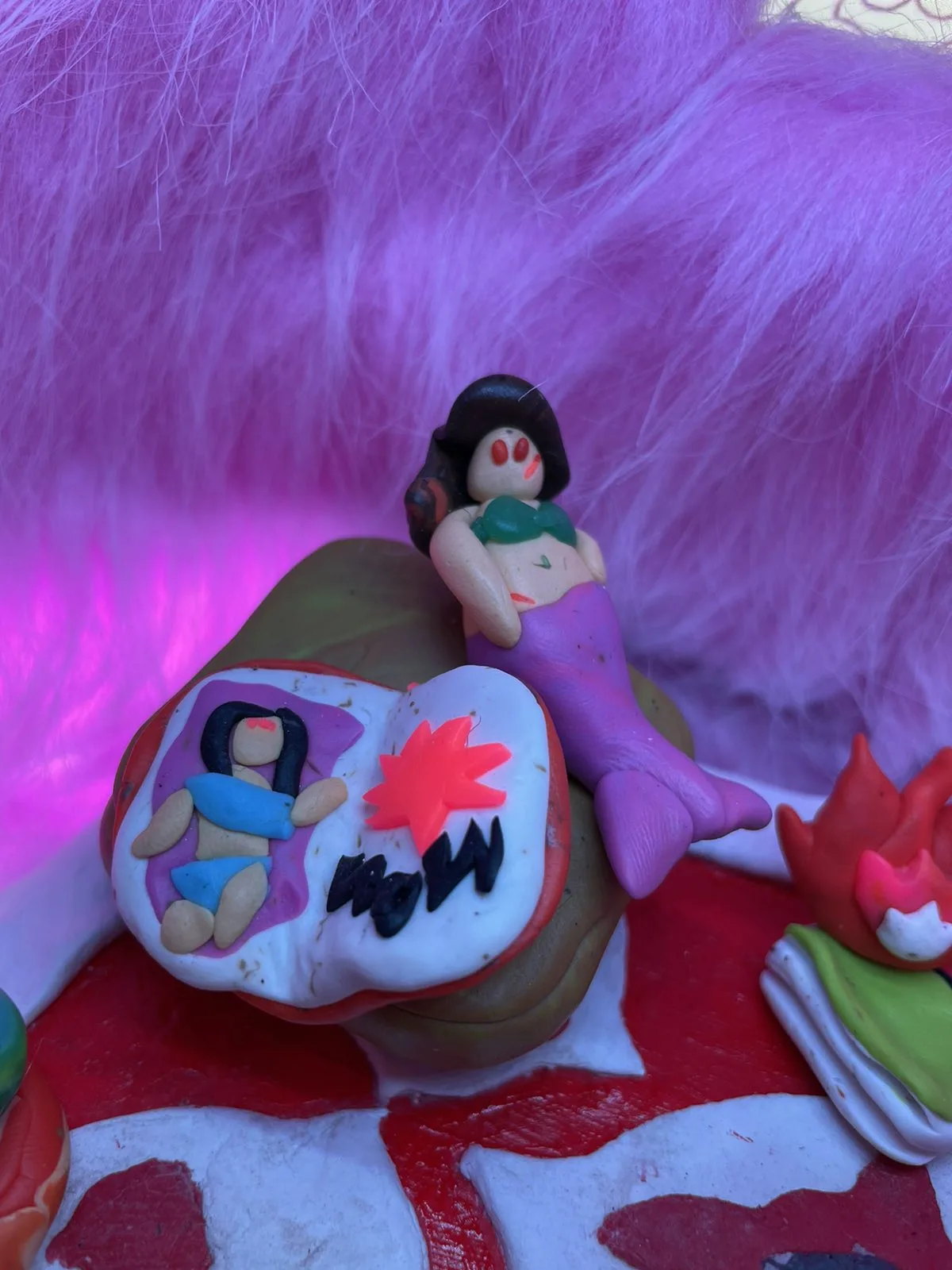 Close-up of a clay mermaid figure reclining against a patterned cushion, under purple light