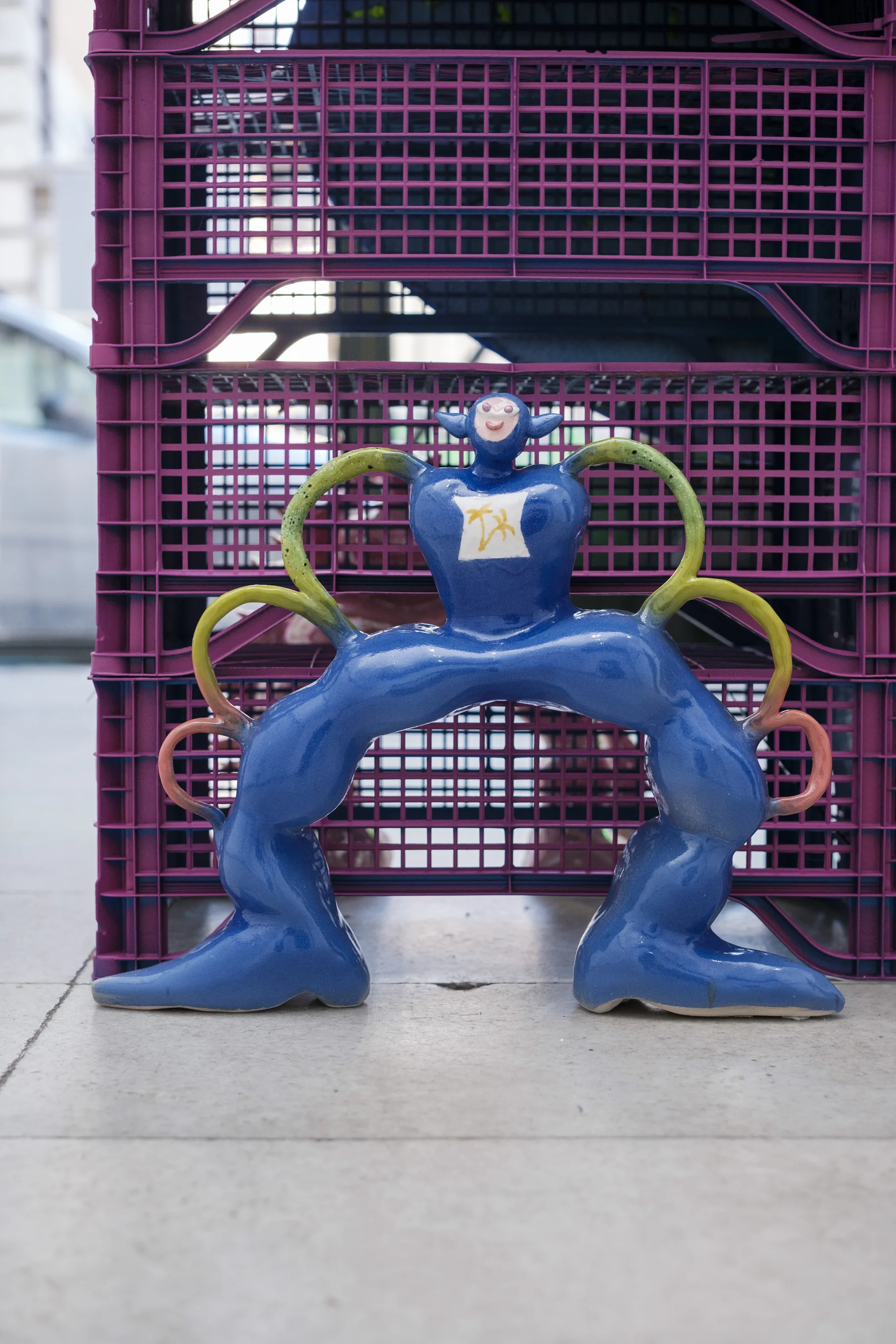 Blue glazed arch-shaped figure with looping arms, standing in front of purple crates