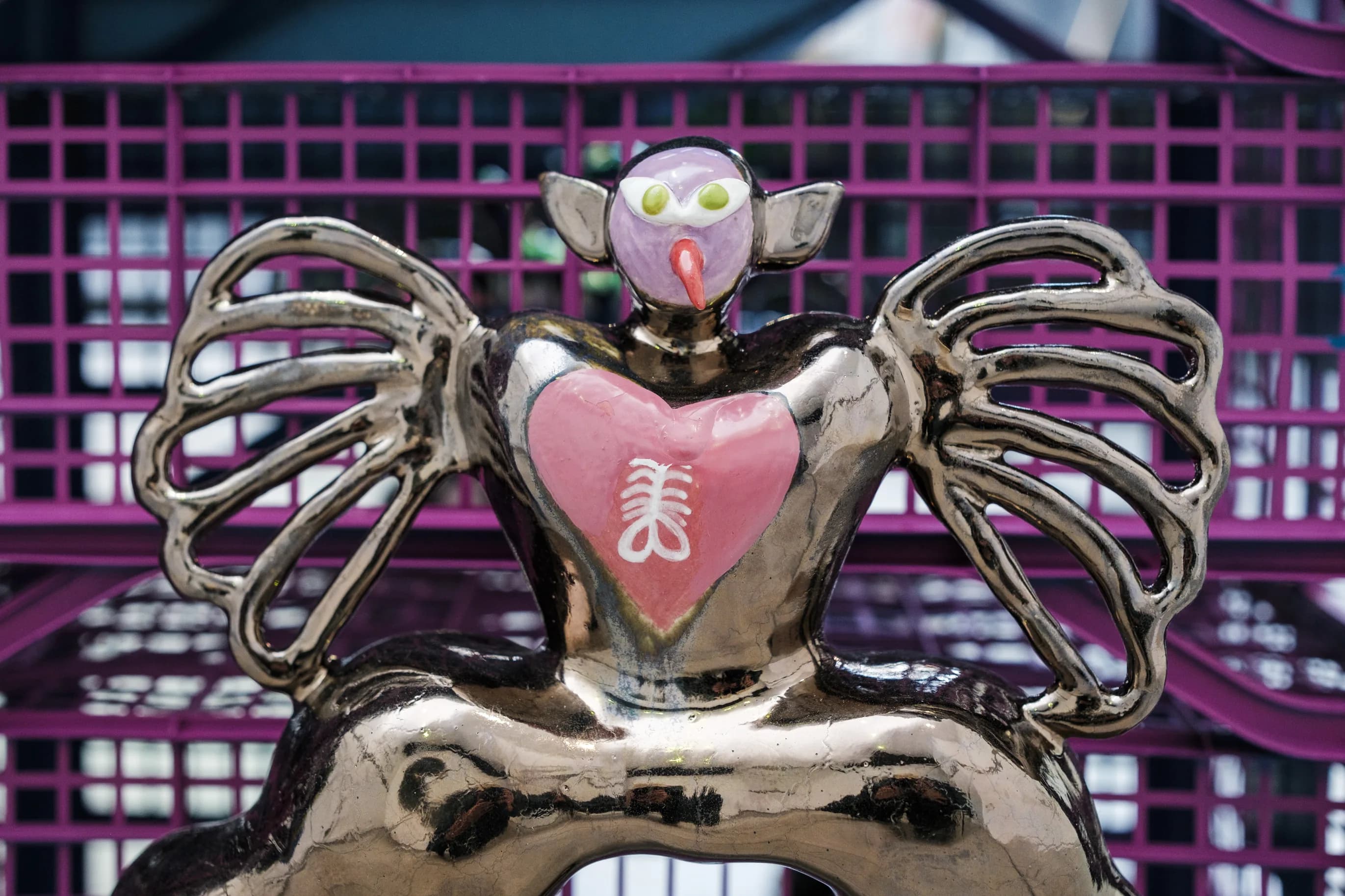 Detail of a bronze-glazed winged figure with a pink heart, in front of purple crates