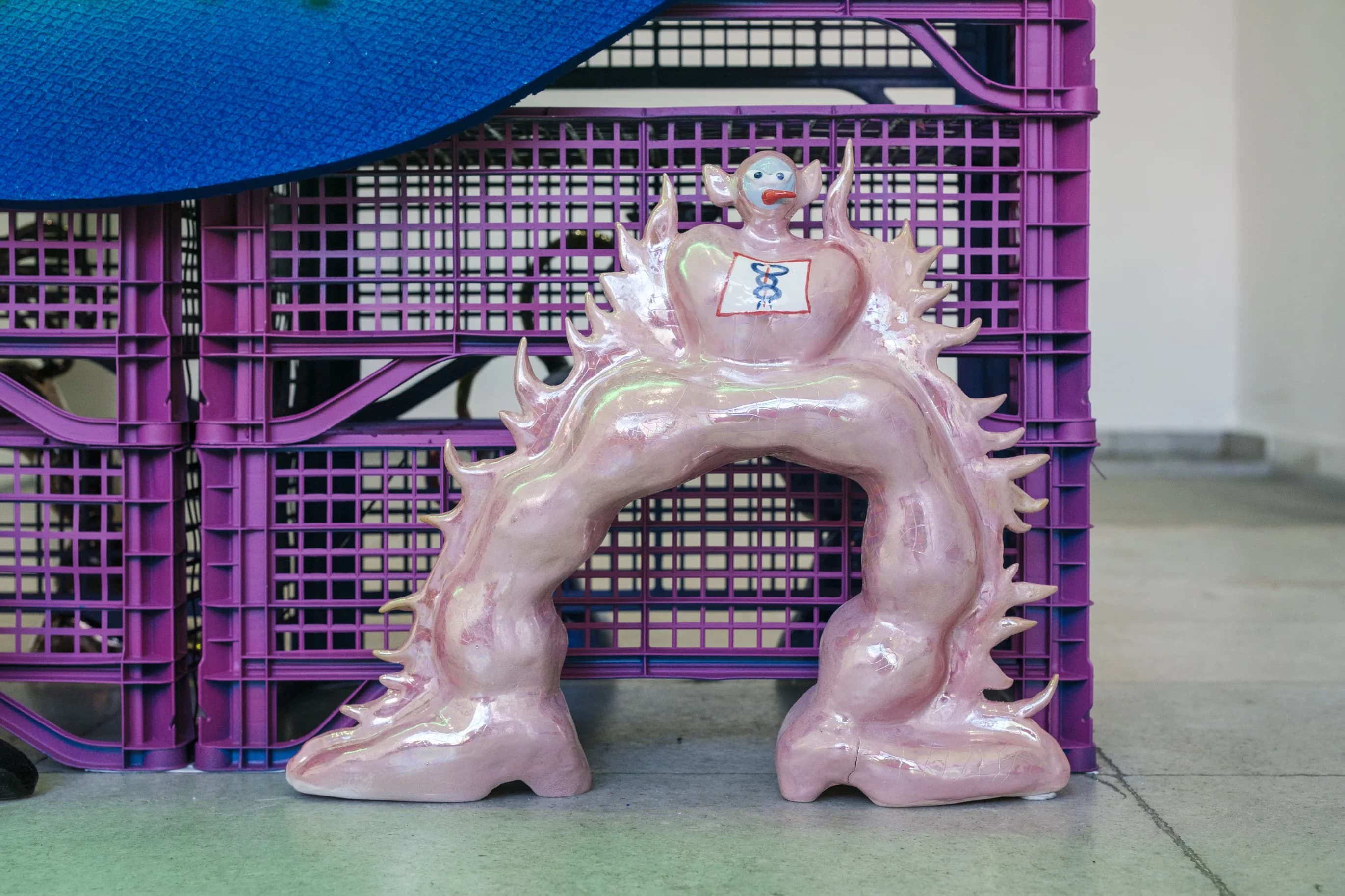 Pink glazed arch-shaped figure covered in spikes, in front of purple crates