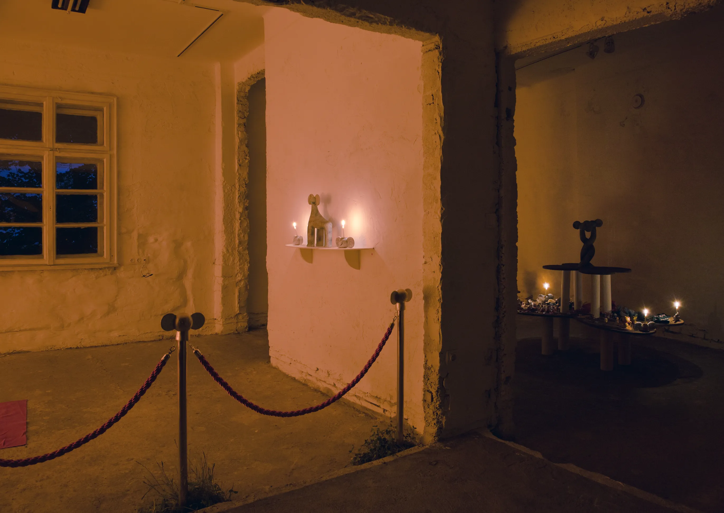 Dusk view of a candlelit shelf sculpture in an alcove, seen past a velvet rope barrier