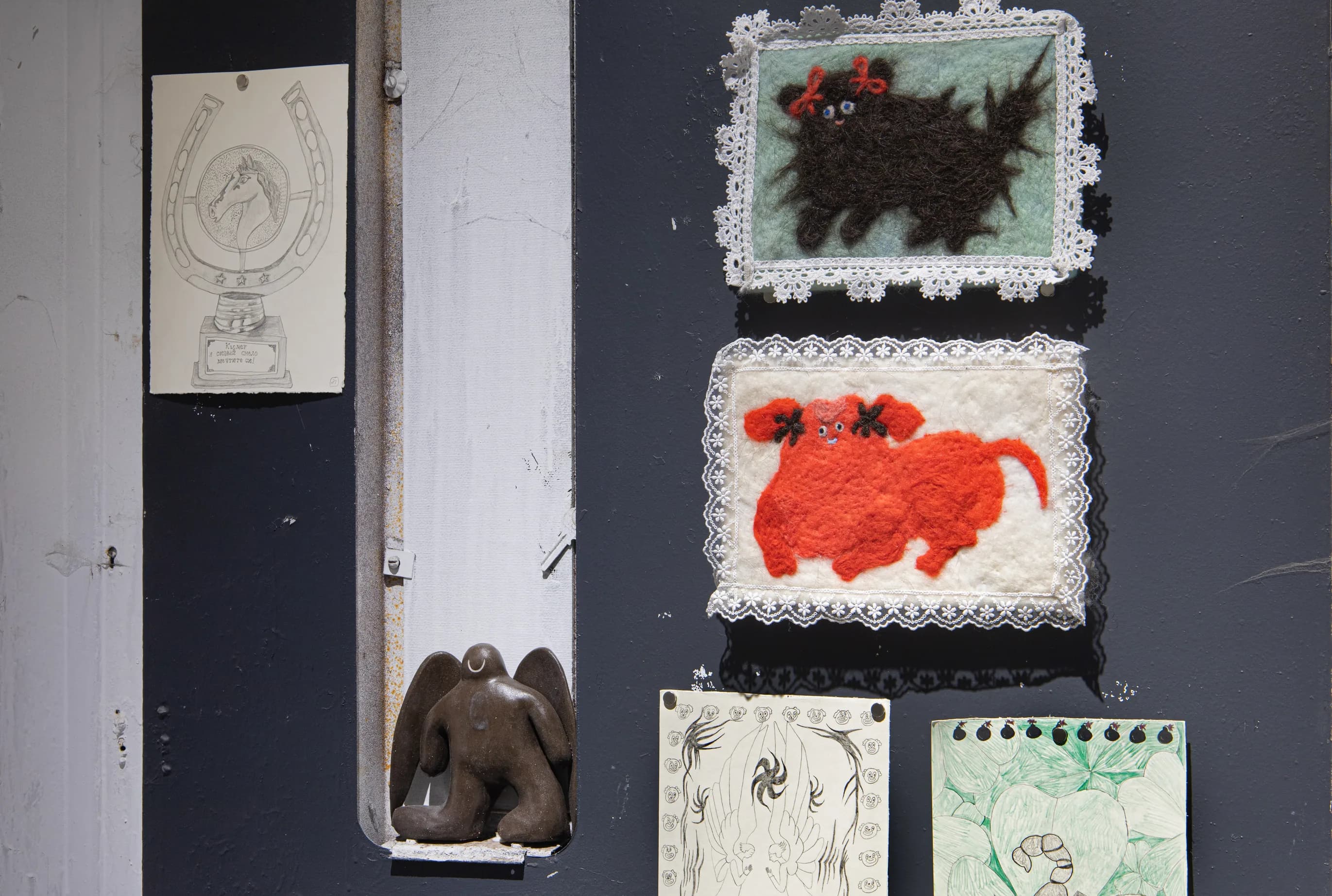 Felted animal pictures in lace frames and small drawings on a dark wall, with a dark winged ceramic figure on a ledge