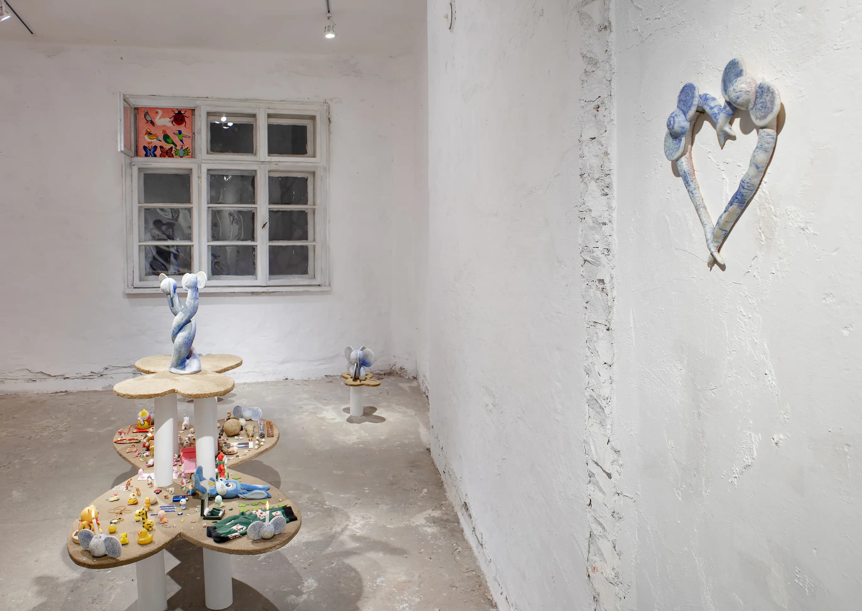 Gallery corner with a heart-shaped wall sculpture, a twisted ceramic figure on a pedestal, and an elephant-eared figurine