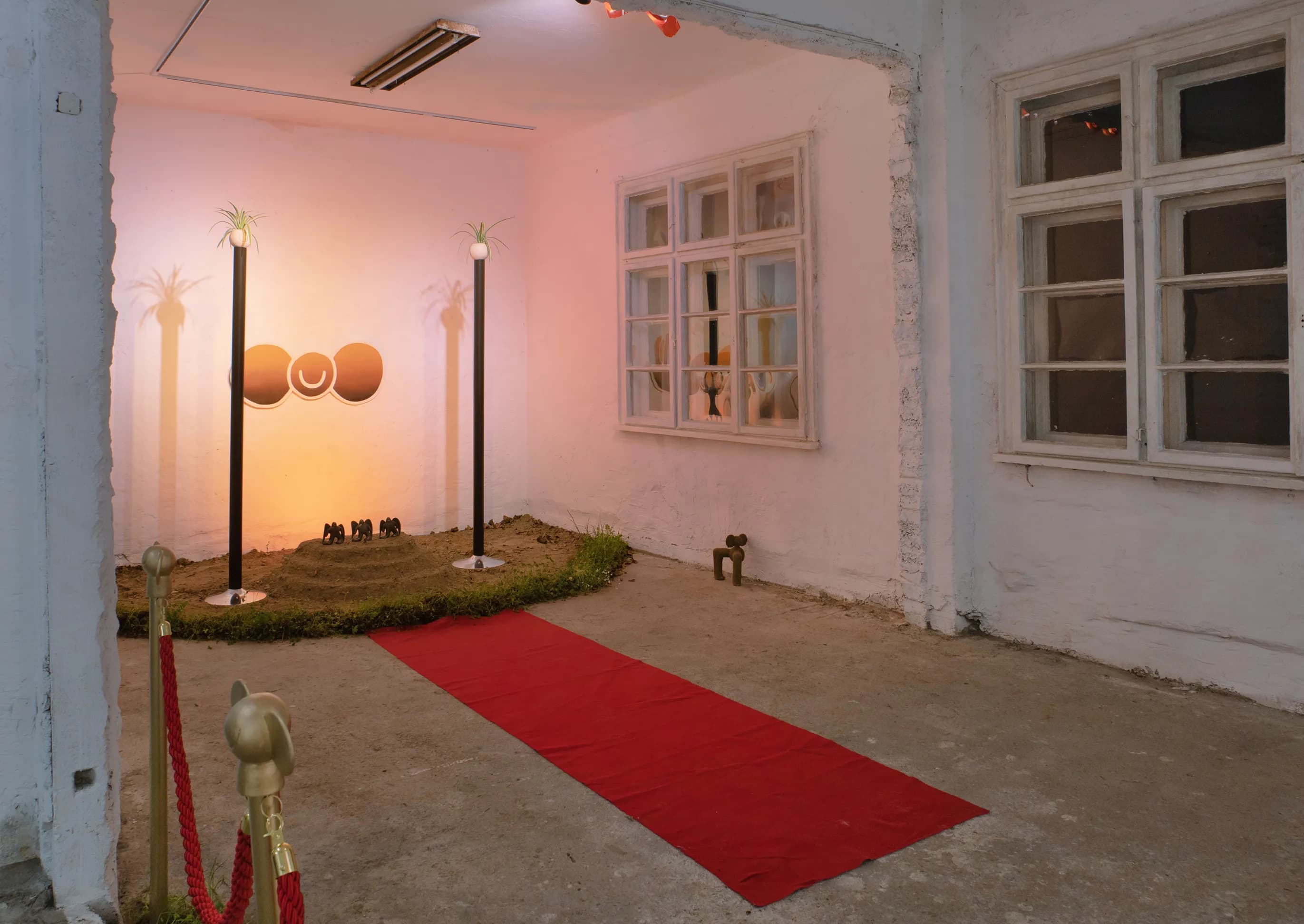 Installation view with a red carpet leading past velvet ropes toward a glowing pink backdrop and a mound with three winged figurines